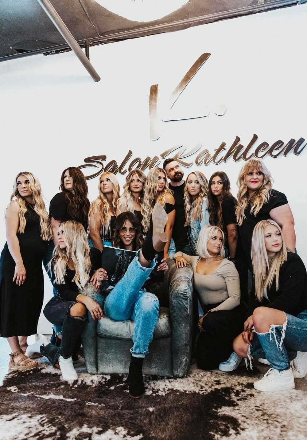 Salon Kathleen's full team posing in front of the salon's logo-adorned wall