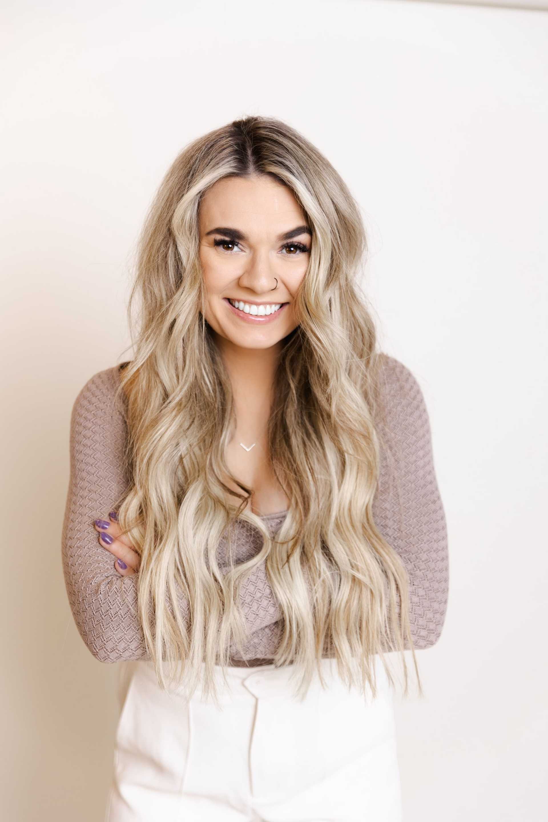 Smiling woman with long blonde hair, wearing a taupe sweater and white pants against a white background.