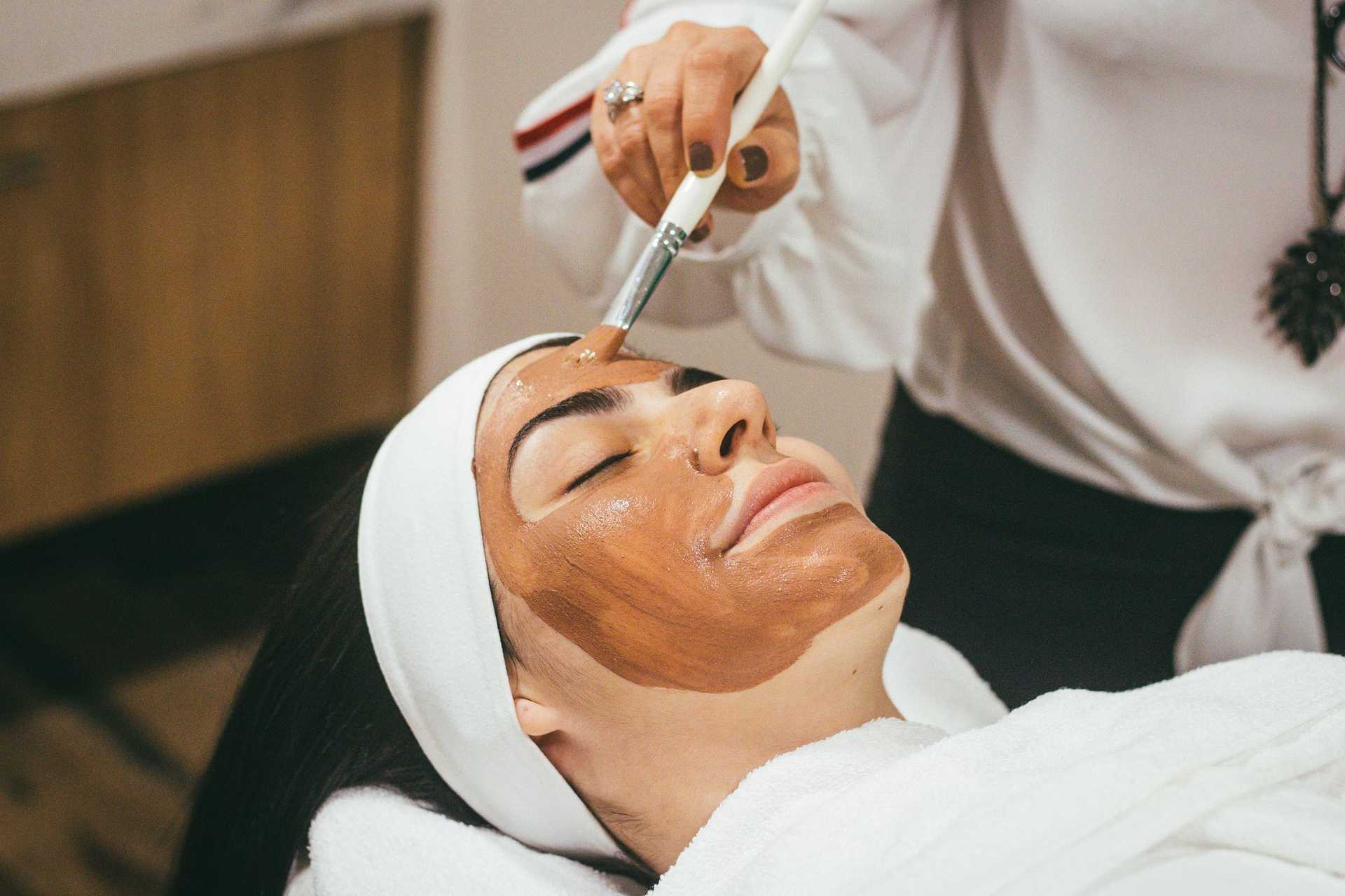An aesthetician applies a facial mask to a relaxed client's skin. The client lies back with eyes closed and hair pulled away from her face, as the aesthetician uses a brush to gently paint a facial mask onto her skin.