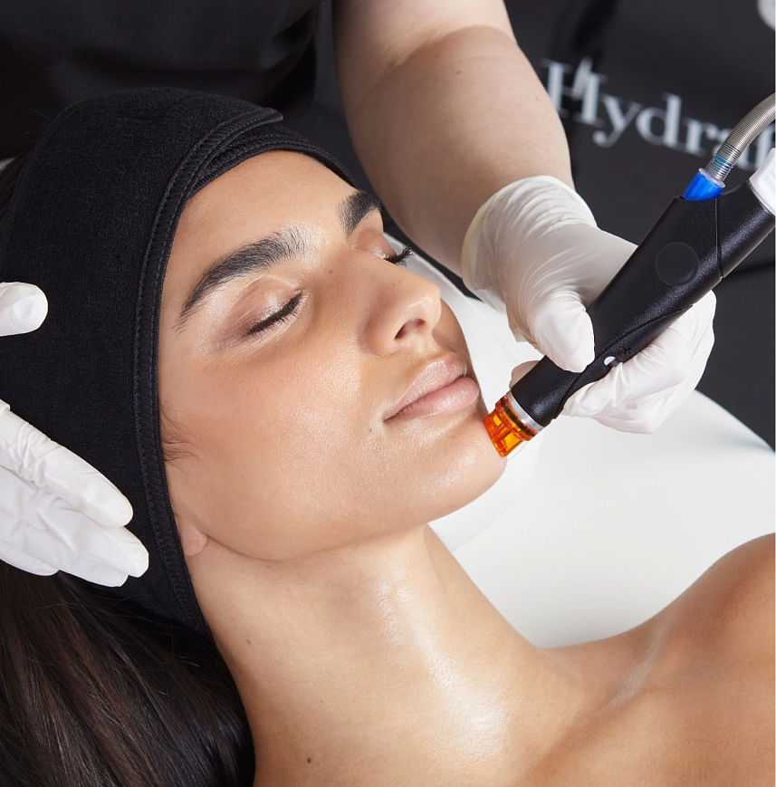 Close-up of Salon Kathleen An aesthetician performing a HydraFacial treatment on the face of a client. The image shows the aesthetician holding the HydraFacial machine tip just above the client's skin. The tip applies gentle suction and serums to deep clean and infuse the skin.