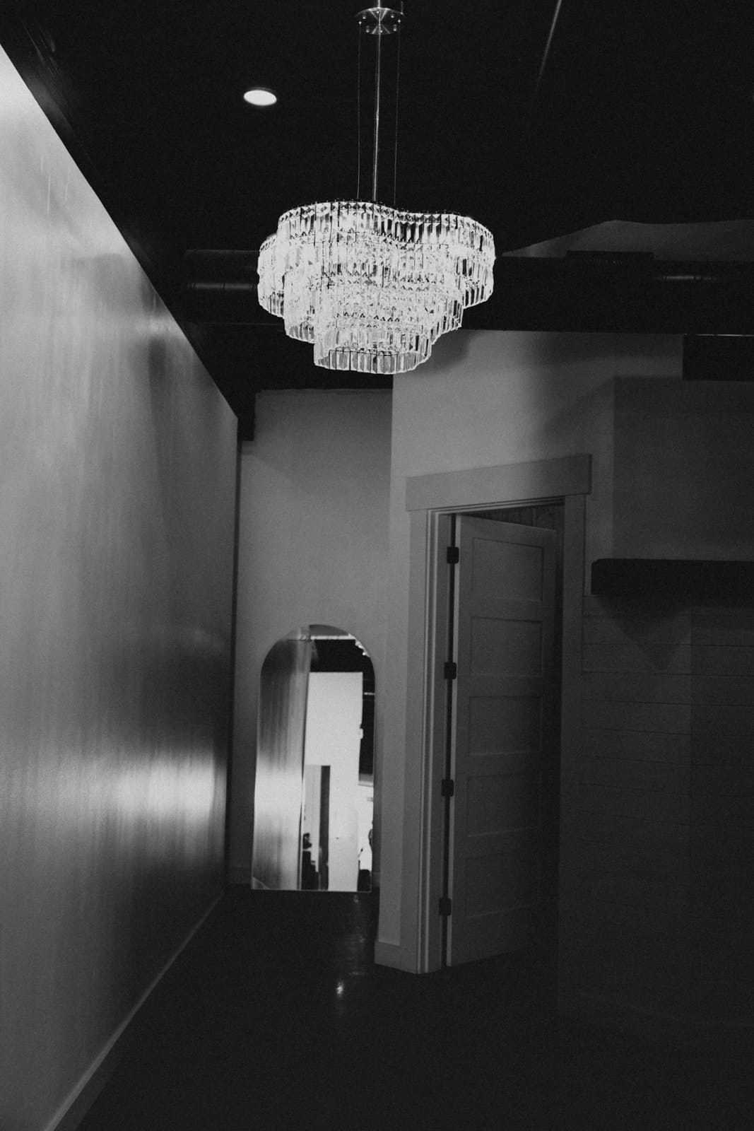 Dimly lit hallway with a sparkling chandelier and an open door leading to a bright room.