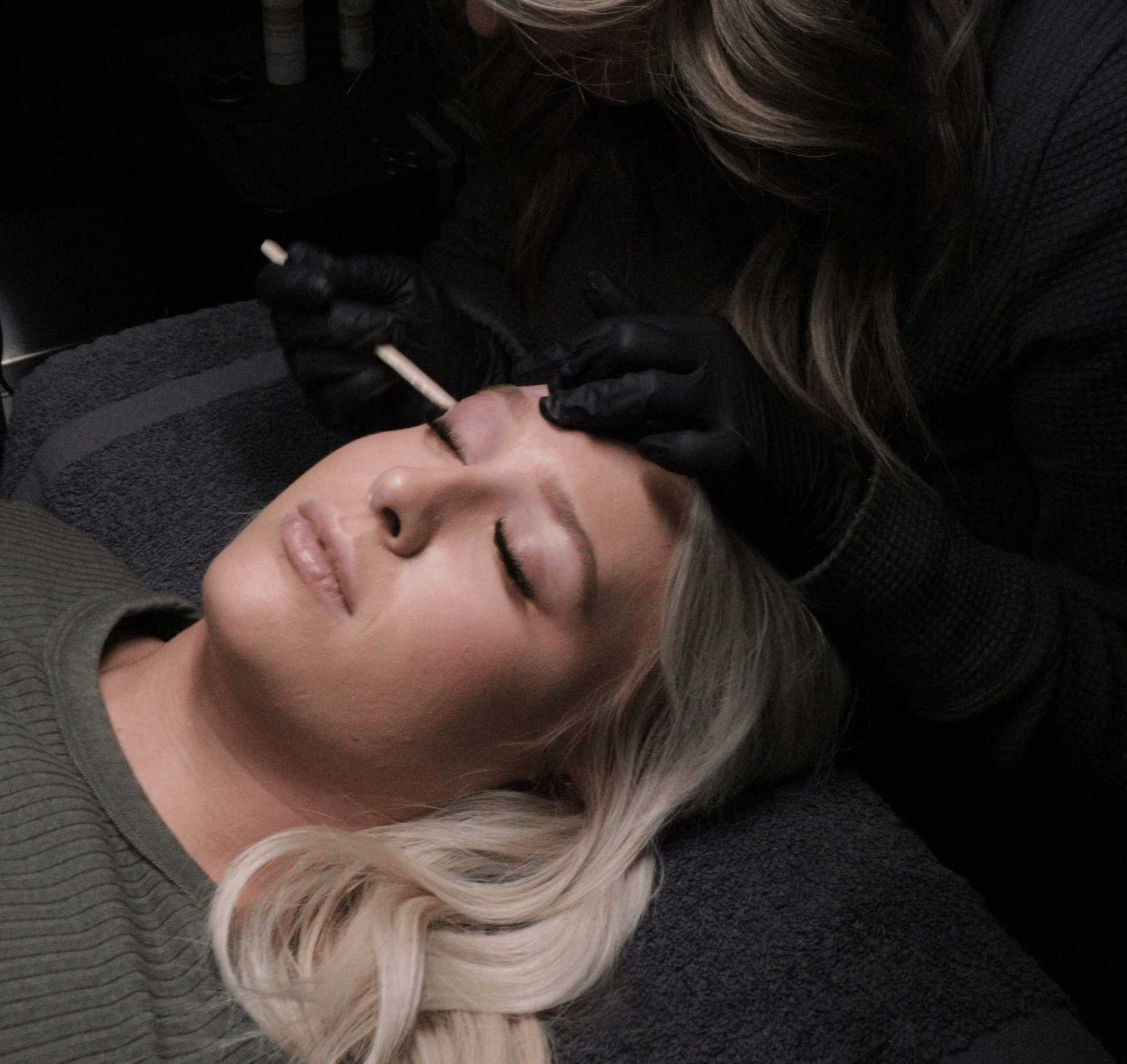 Aesthetician carefully shaping a client's eyebrows during a brow waxing service at Salon Kathleen. The client is reclining with eyes closed as the aesthetician meticulously sculpts and waxes for a polished end result.