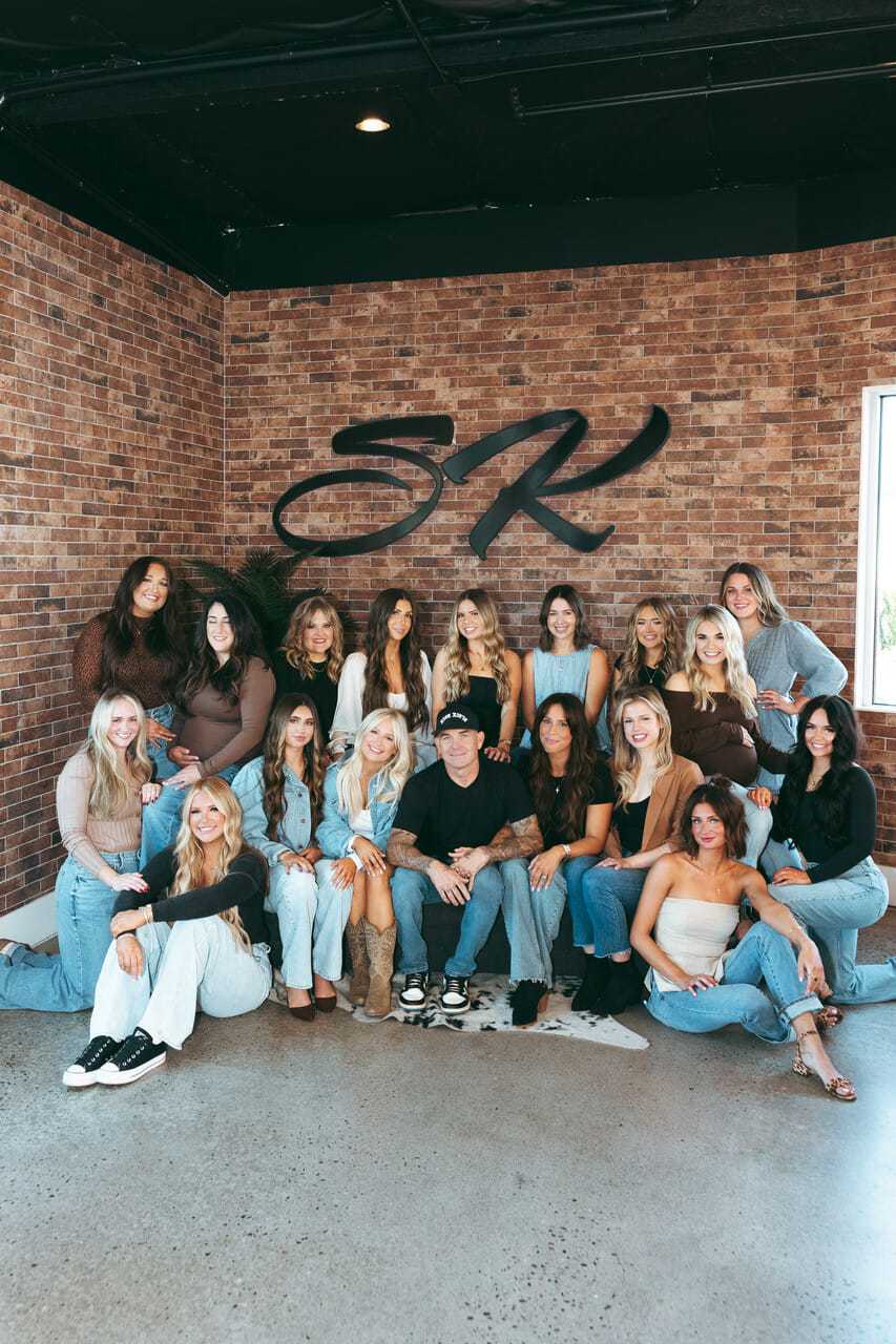 Group of people posing in a studio with a brick wall background and a large logo.