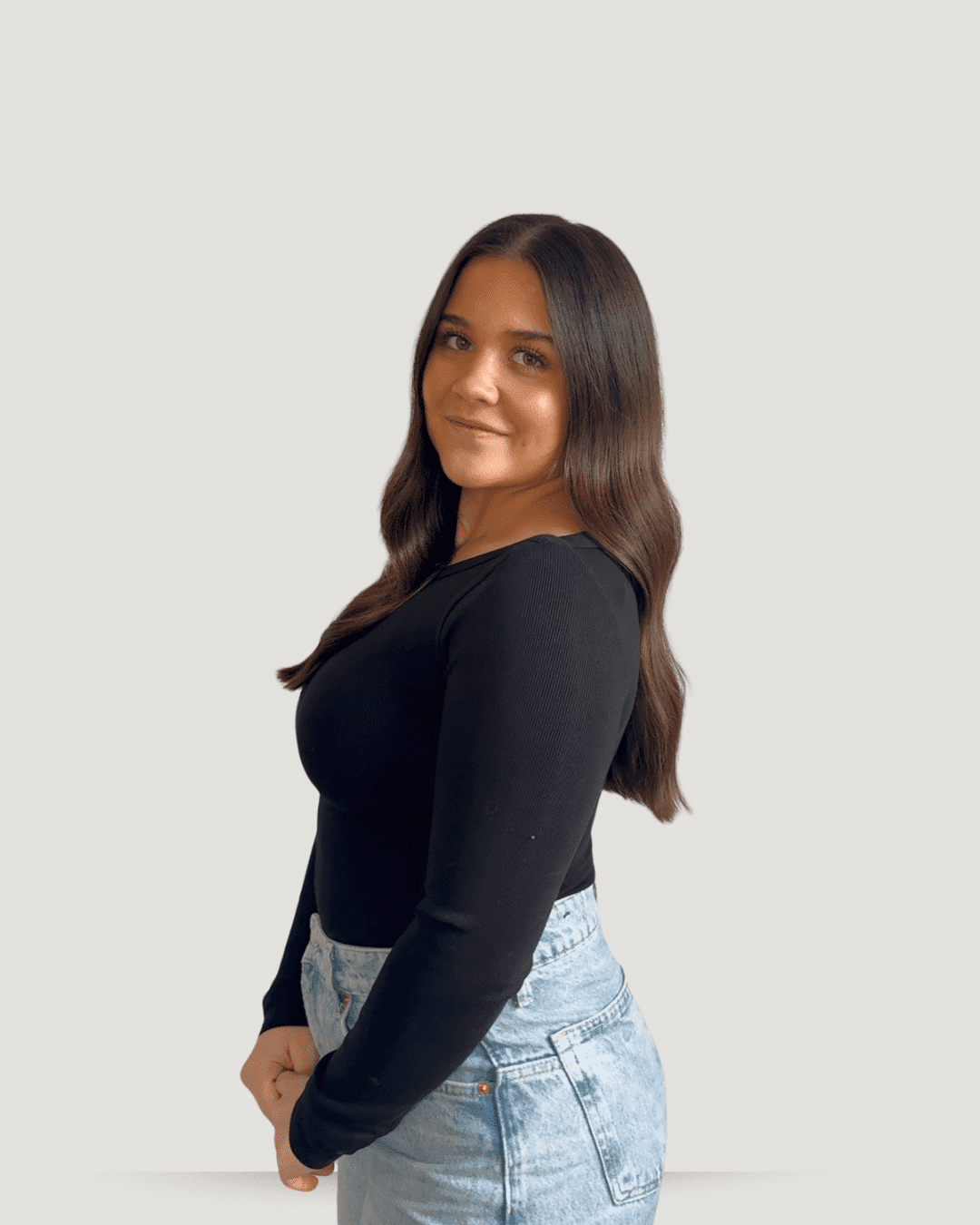Young woman with long hair wearing a black top and jeans against a plain background.