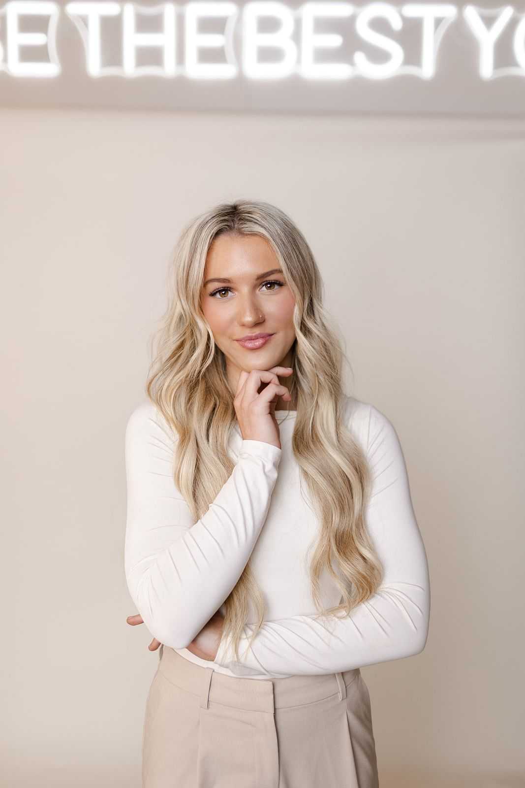 Blonde woman in white top posing with hand on chin under neon sign reading "BE THE BEST YOU".