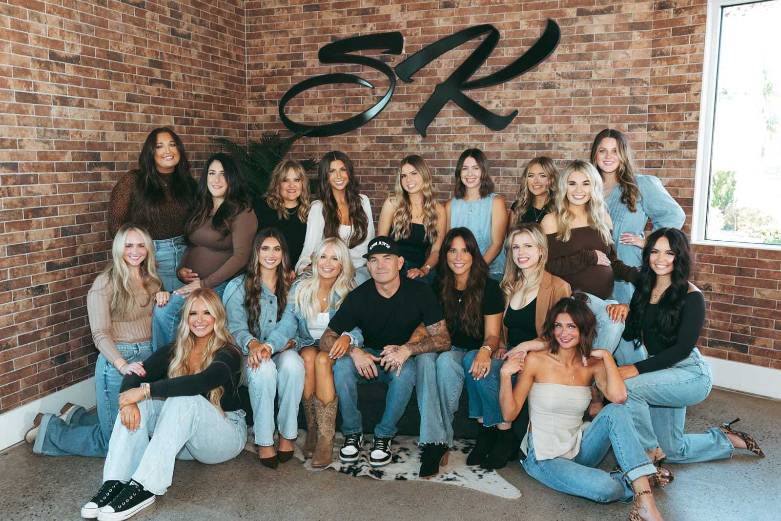 Group photo of 18 smiling people in casual attire against a brick wall with a logo.