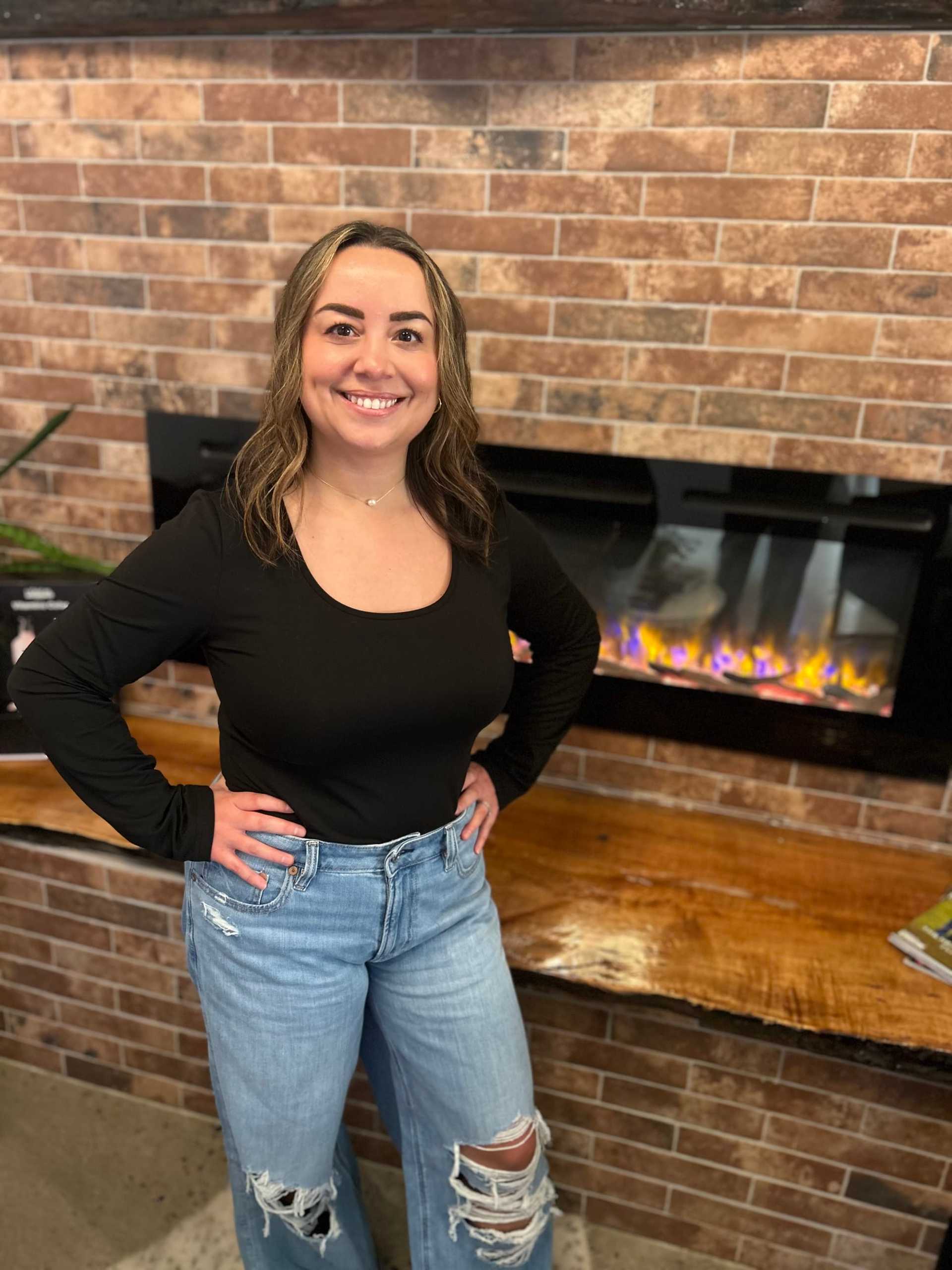 Woman smiling by a brick fireplace, wearing a black top and ripped jeans.