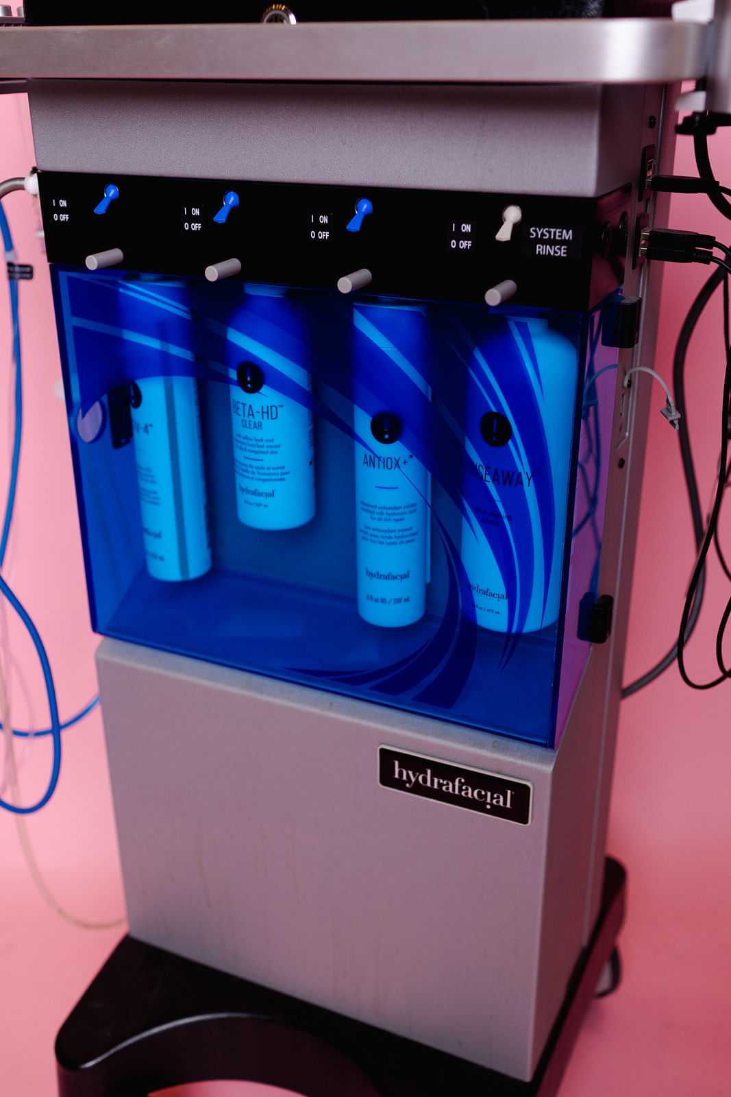 Hydrafacial machine with skincare product bottles and control buttons visible against a pink backdrop.