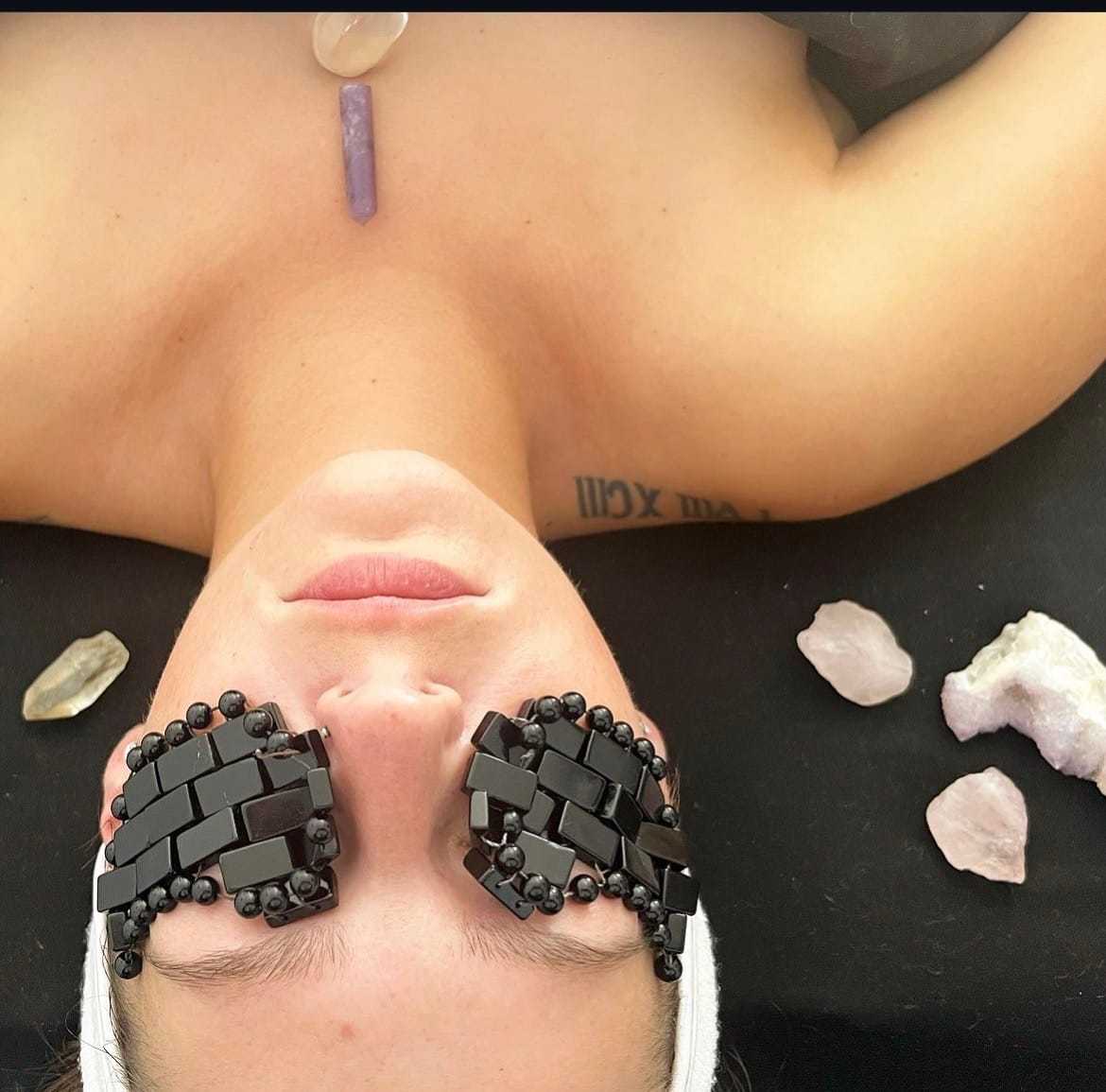 Person with gemstone eye masks lies surrounded by crystals on a black surface.